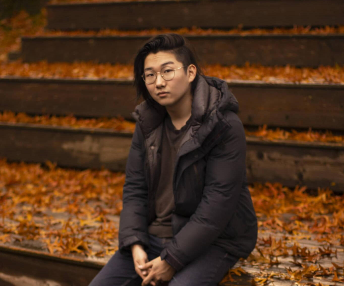 Portrait of Max Kim wearing a navy blue puffer jacket, sitting outdoors with damp wooden steps and scattered orange fallen leaves softly blurred in the background, evoking a calm autumn atmosphere.